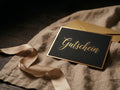 Eine Nahaufnahme einer luxuriösen, gold geränderten schwarzen Gutscheinkarte, die auf einem strukturierten beige-grauen Leinentuch auf einem dunklen Holztisch liegt. Der Text 'Gutschein' ist in einer eleganten, goldenen Schreibschrift auf der Karte gedruckt. Ein goldener Umschlag ist teilweise hinter der Karte sichtbar, und ein champagnerfarbenes Satinband schlängelt sich im Vordergrund. Die Beleuchtung ist warm und atmosphärisch.